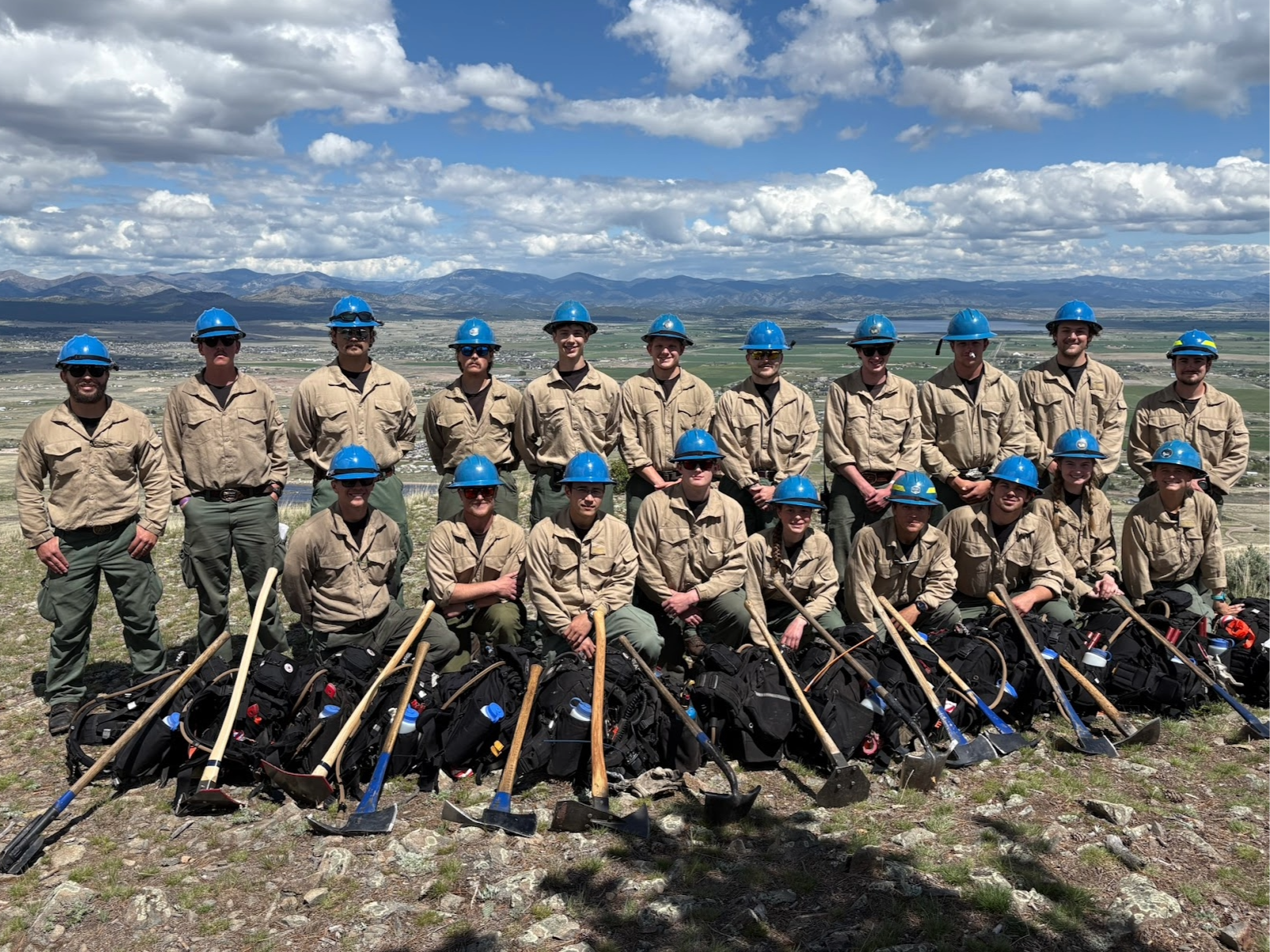 Outdoor group portrait of approximately twenty wildland firefighters wearing matching tan long-sleeve shirts, green pants, and blue hard hats, standing and kneeling in two rows on a rocky hillside with their faces intentionally blurred; a row of hand tools and black equipment packs is laid out in front of them, and an expansive valley with fields, a body of water, and distant mountain ranges appears under a partly cloudy blue sky in the background.