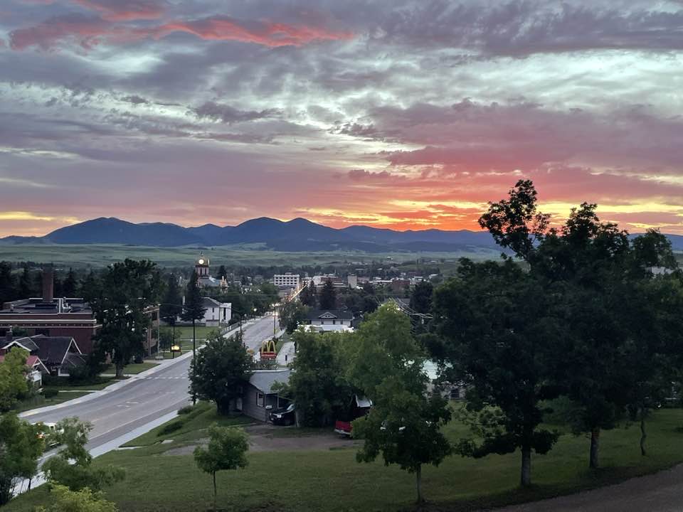 A small street in Lewistown leads toward distant rounded mountains under a dramatic pink and orange sunset sky, with scattered trees and houses in the foreground.