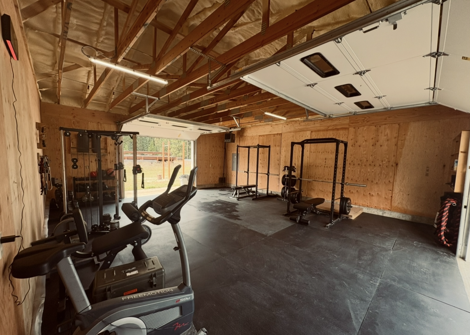 Small weight room inside a wooden-walled garage with an open door, showing a power rack with barbell, cable machine, dumbbell racks, exercise bike, and fields and trees visible outside.