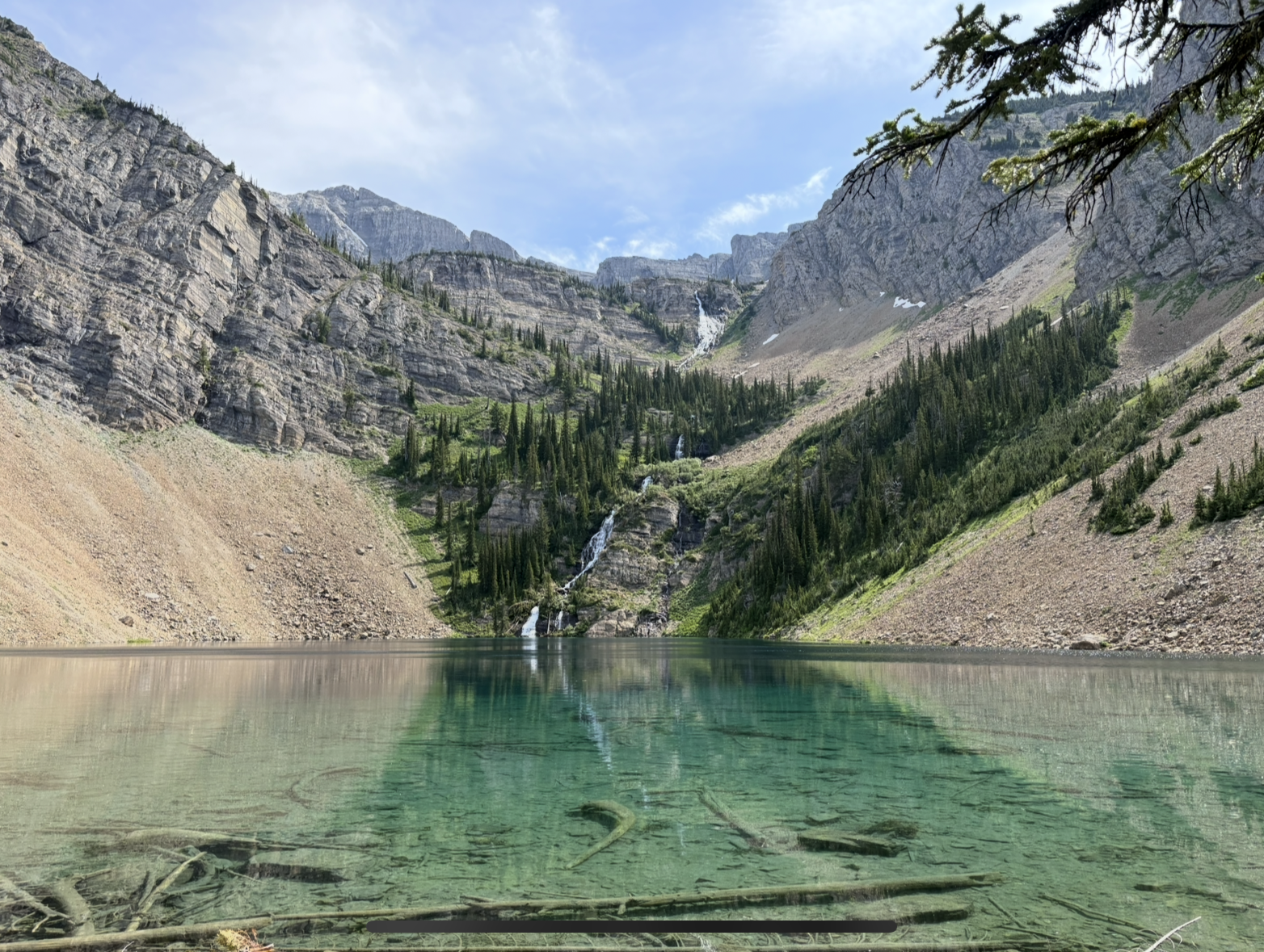 Clear alpine lake with submerged logs in the foreground, steep rocky slopes on both sides, and a cascading waterfall descending through green trees from high cliffs under a partly cloudy sky.
