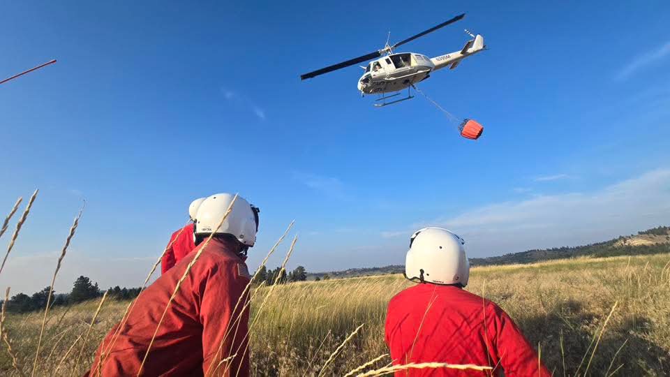 Firefighters watch hovering helicopter carrying orange water bucket during training.