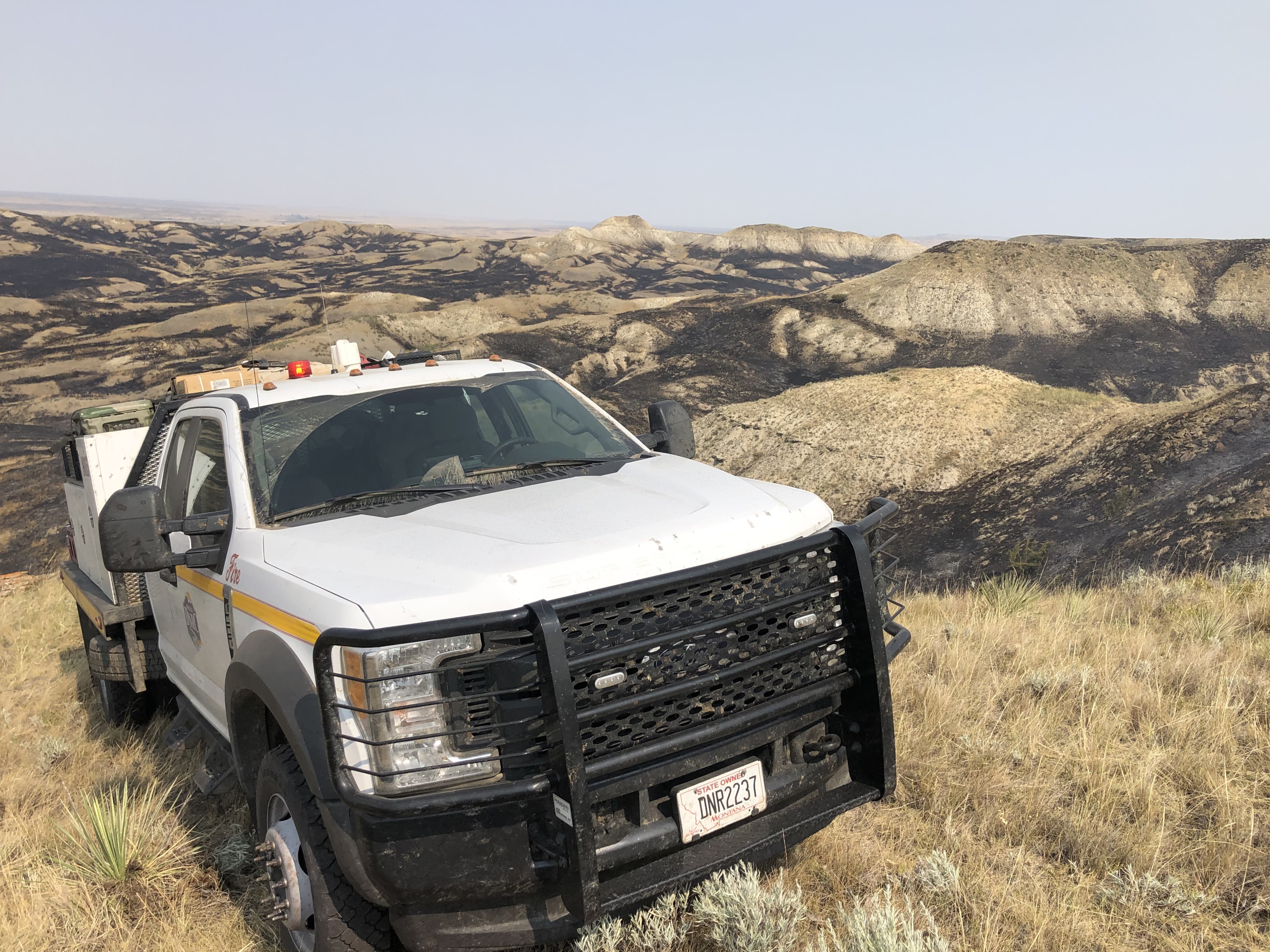 A white DNRC wildland fire pickup truck is parked on a grassy hillside above rolling, partially blackened hills that show recent wildfire burn patterns in rural Montana.