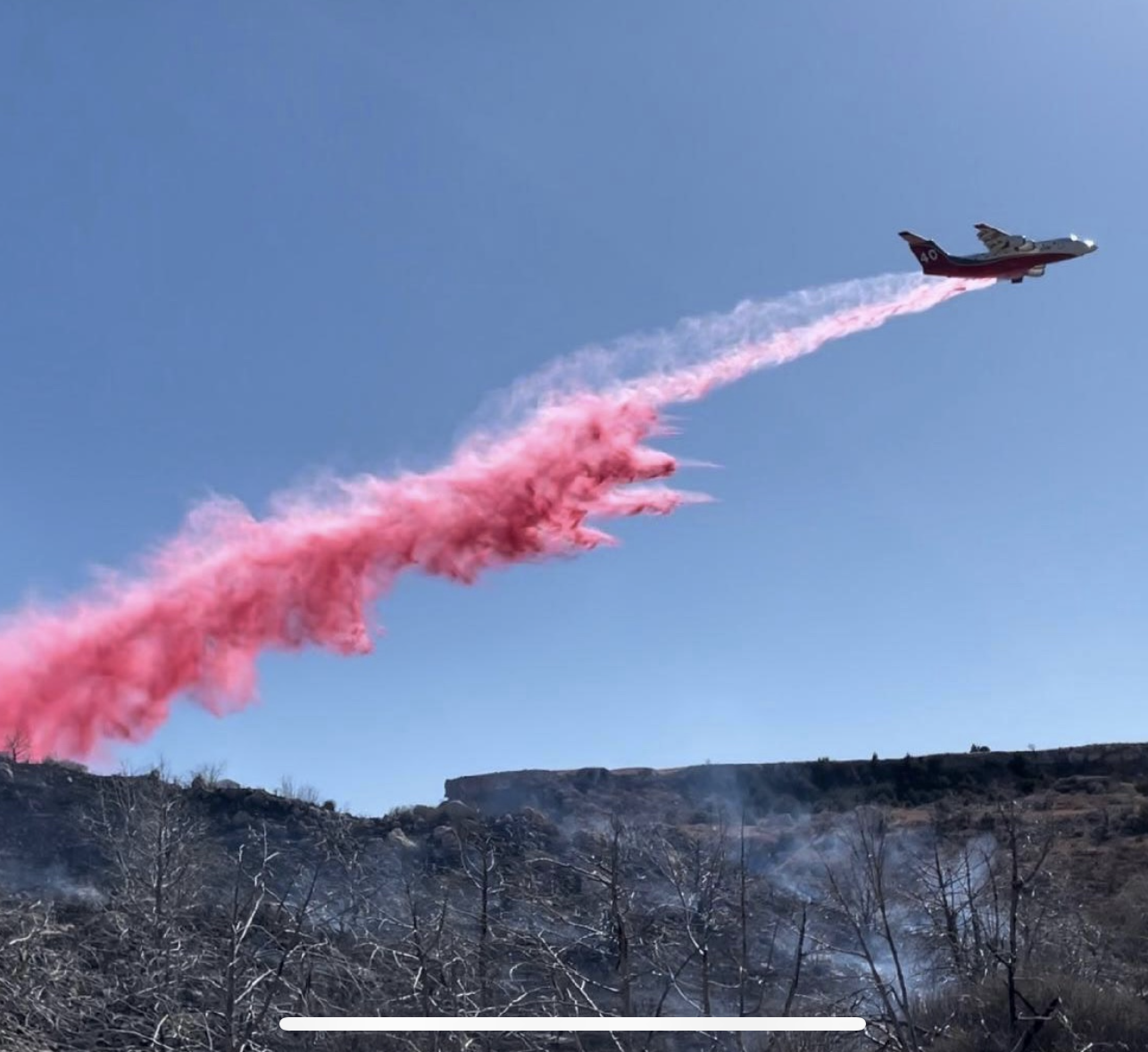 Low-flying firefighting airplane under a clear blue sky releasing a long red plume of retardant over a burned hillside with blackened trees and sparse vegetation.